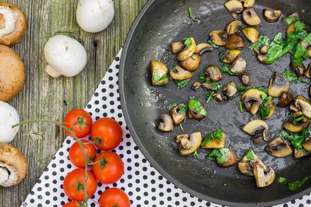 Cooked mushrooms in a pan with tomatoes and raw mushrooms spread out next to it