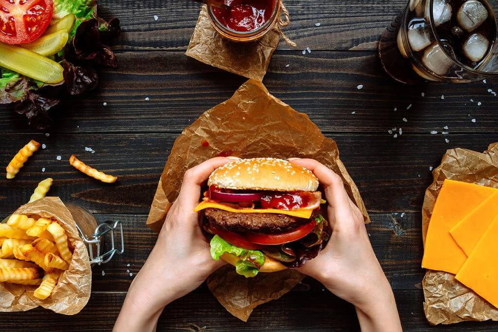 Person holding burger surrounded by cheese slices, soda, ketchup, veggies, and fries