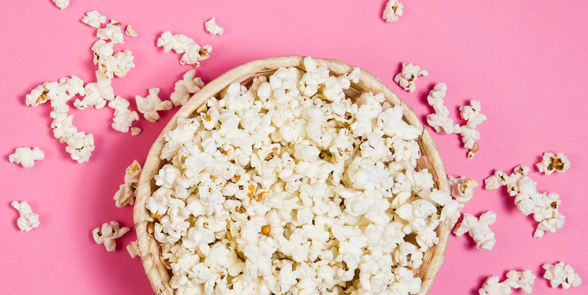 Top-down view of popcorn in a woven basket spilling over onto a pink background