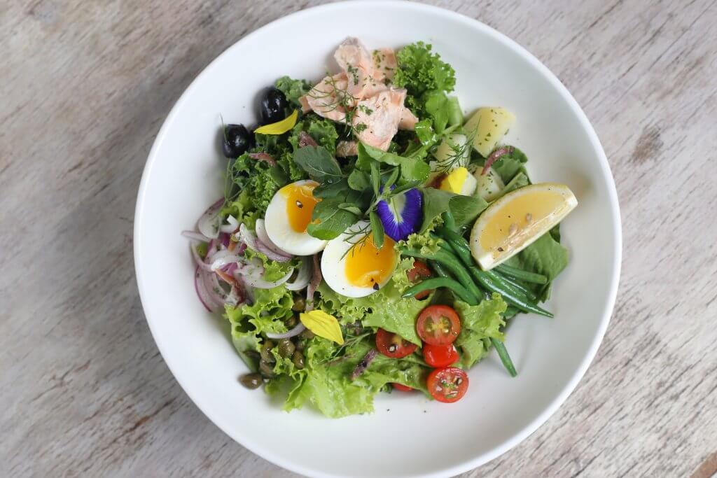 Niçoise salad in a white dish. Two half boiled eggs, blanched green beans, olives, cherry tomatoes and poached salmon arranged nicely on top of mixed greens.