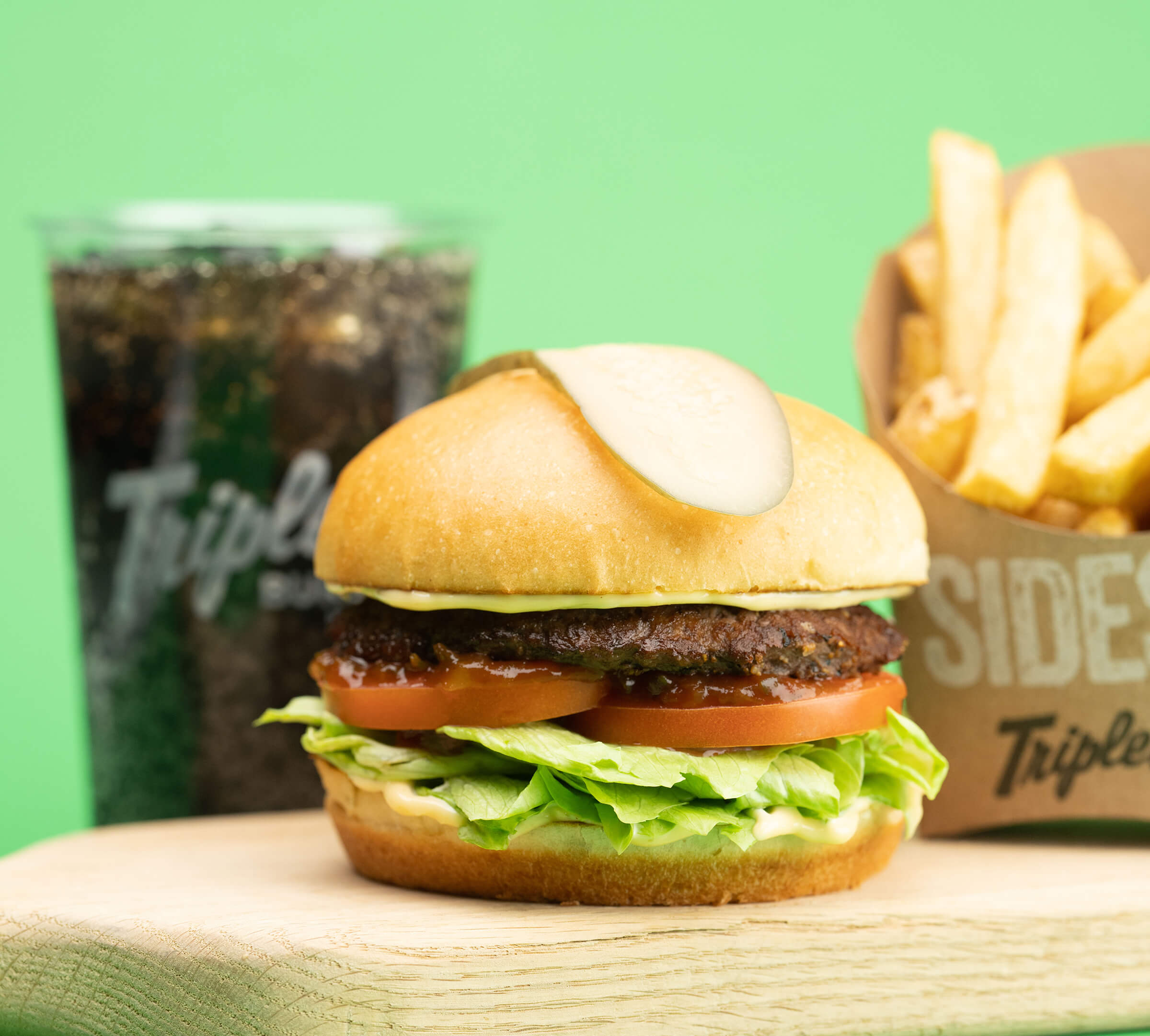 A fully dressed Triple O's burger with an iconic pickle on the bun, fries and a soft drink on a light green background.